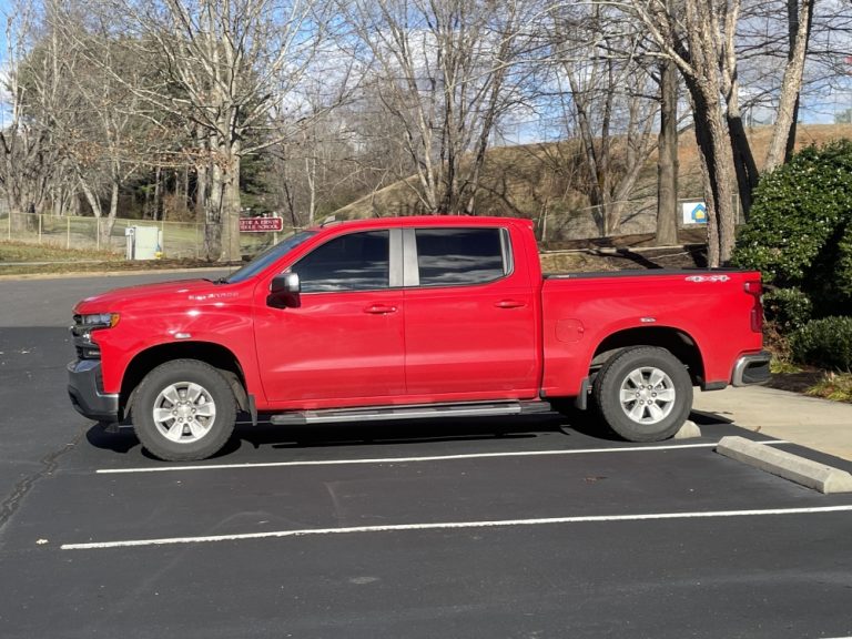 A red pickup truck is parked in an empty parking lot. Bare trees and some greenery are visible in the background along with a stretch of grass and a few signs. It appears to be a sunny day.