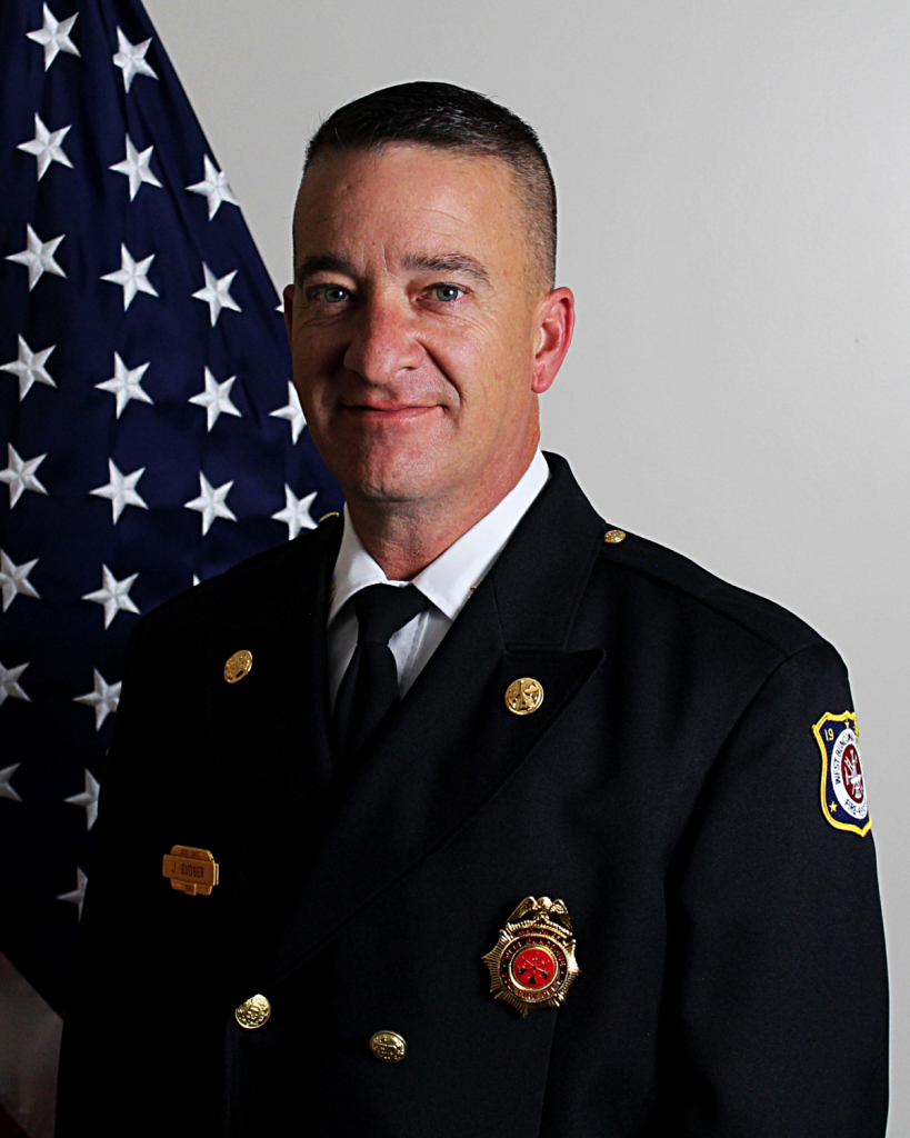 A man in a formal firefighter uniform poses in front of the American flag. He has short hair and a slight smile. The uniform has various insignias and a badge. The background is plain, accentuating the flag and the uniform details.