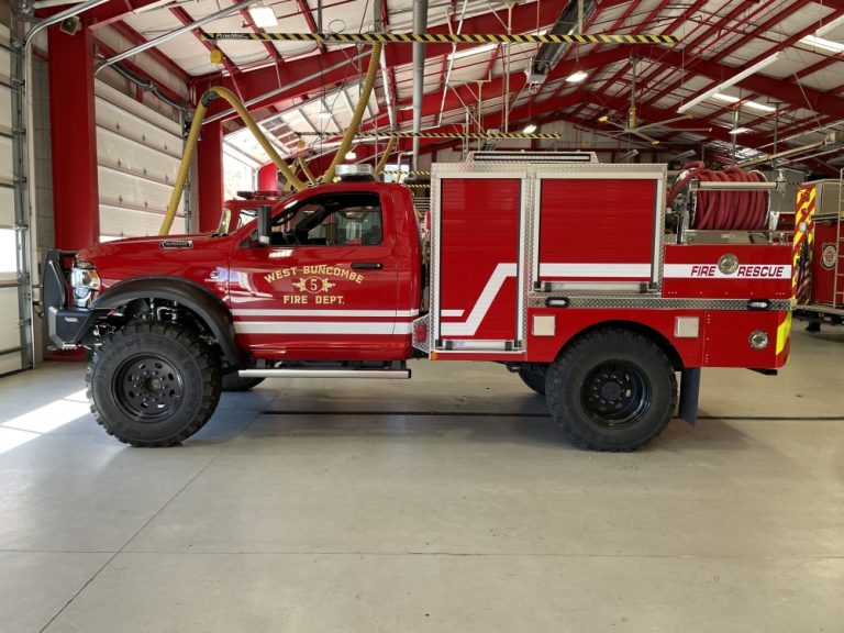 A red fire and rescue truck from the West Buncombe Fire Department is parked inside a fire station. The truck features large off-road tires, equipment compartments, and a roll of hose at the back. The station has red and white steel beams and overhead doors.