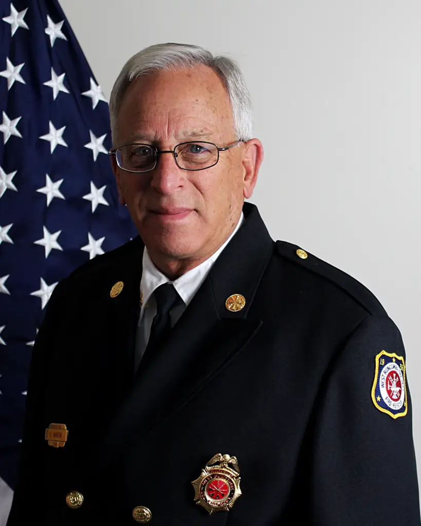 A senior man in a formal firefighter uniform, featuring multiple pins and badges, stands in front of a U.S. flag. He has short gray hair, glasses, and a serious expression. The uniform displays insignia and an embroidered patch on the sleeve.