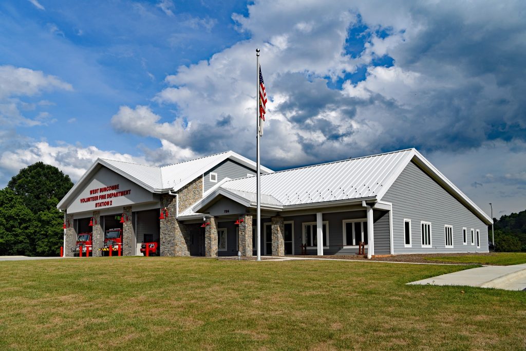A modern fire station with gray siding and a white metal roof under a partly cloudy sky. An American flag flies on a tall flagpole in front of the building. Fire trucks are visible in the open garage bays. A neatly mowed lawn surrounds the station.