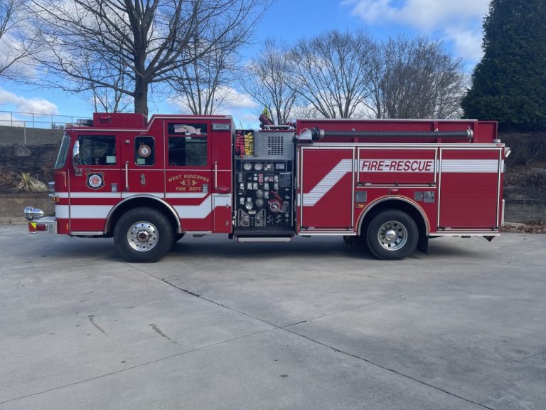 A red fire-rescue truck from the West Mead Township Volunteer Fire Department, featuring white stripes and various compartments, is parked on a concrete surface. The background includes bare trees and a clear, slightly cloudy blue sky.