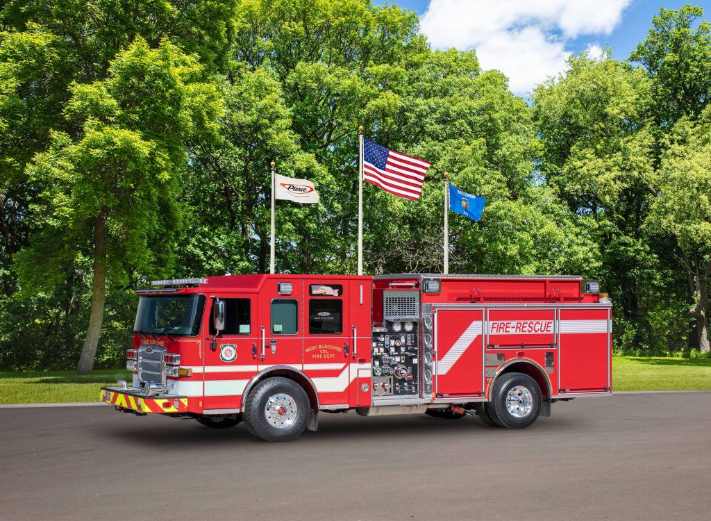 A red and white fire truck with "Fire-Rescue" written on its side is parked on a paved surface with lush green trees and three flags (one of which is the American flag) in the background. The sky is partly cloudy with patches of blue.