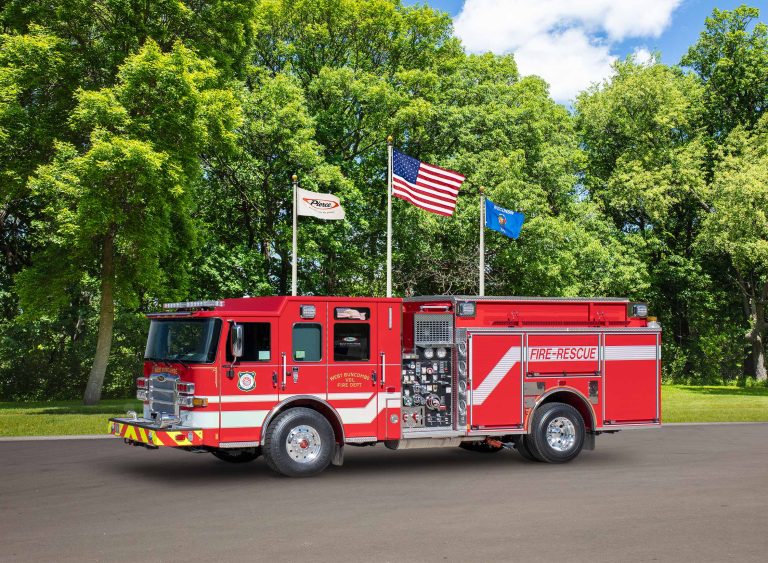 A red and white fire truck with "Fire-Rescue" written on its side is parked on a paved surface with lush green trees and three flags (one of which is the American flag) in the background. The sky is partly cloudy with patches of blue.