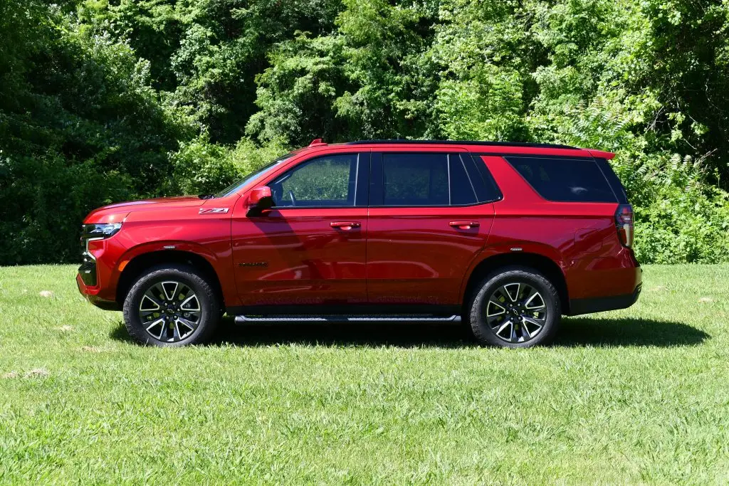 A red SUV is parked on a grassy area with a backdrop of green, leafy trees. The vehicle is shown in profile from the driver's side, displaying its full side view, including the wheels and windows. The SUV has black trim and roof rails.
