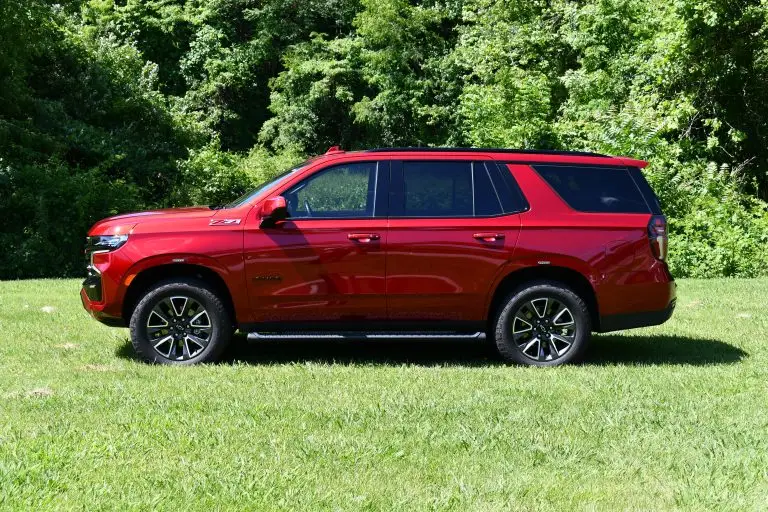 A red SUV is parked on a grassy area with a backdrop of green, leafy trees. The vehicle is shown in profile from the driver's side, displaying its full side view, including the wheels and windows. The SUV has black trim and roof rails.