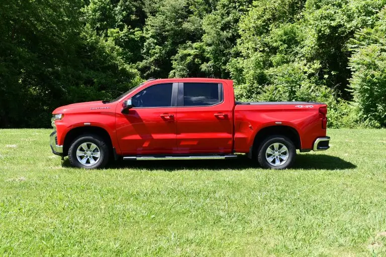 A bright red pickup truck with a double cab is parked on a grassy field with dense trees in the background. The truck has a sleek design with a shiny exterior.