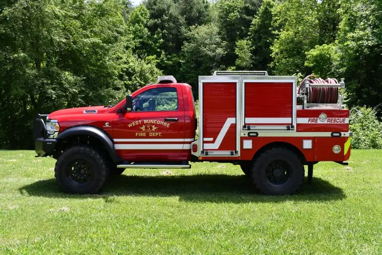 A bright red fire rescue truck from the West Buncombe Fire Department is parked on grass, surrounded by lush green trees. The truck's side compartments and equipment are visible, including hoses on top and department markings on the door.