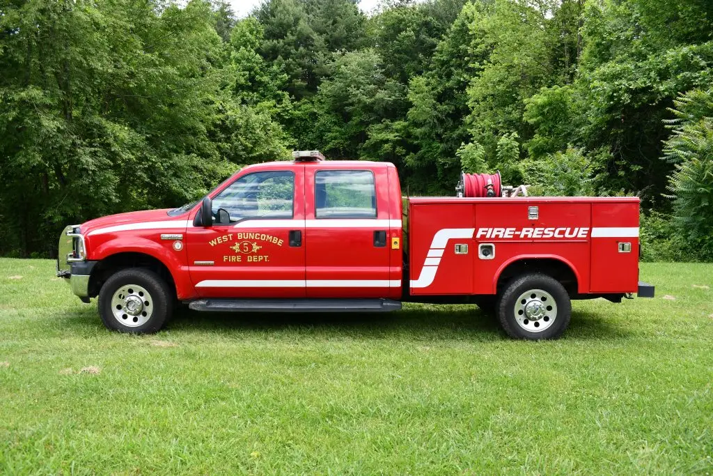 A bright red fire-rescue truck from the West Buncombe Fire Department is parked on green grass with a forested backdrop. The truck features various compartments and equipment, including a coiled fire hose on the rear, and has emergency lights on top.