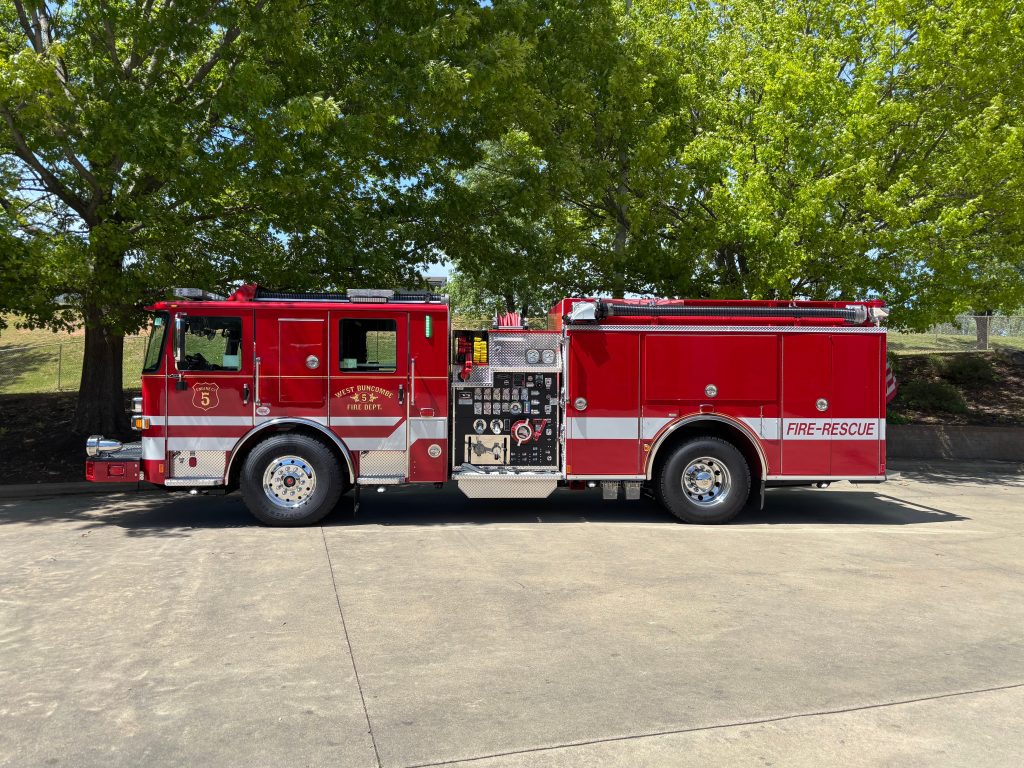 A bright red fire-rescue truck marked "Engine 5" is parked on a paved surface, with green trees in the background on a sunny day.