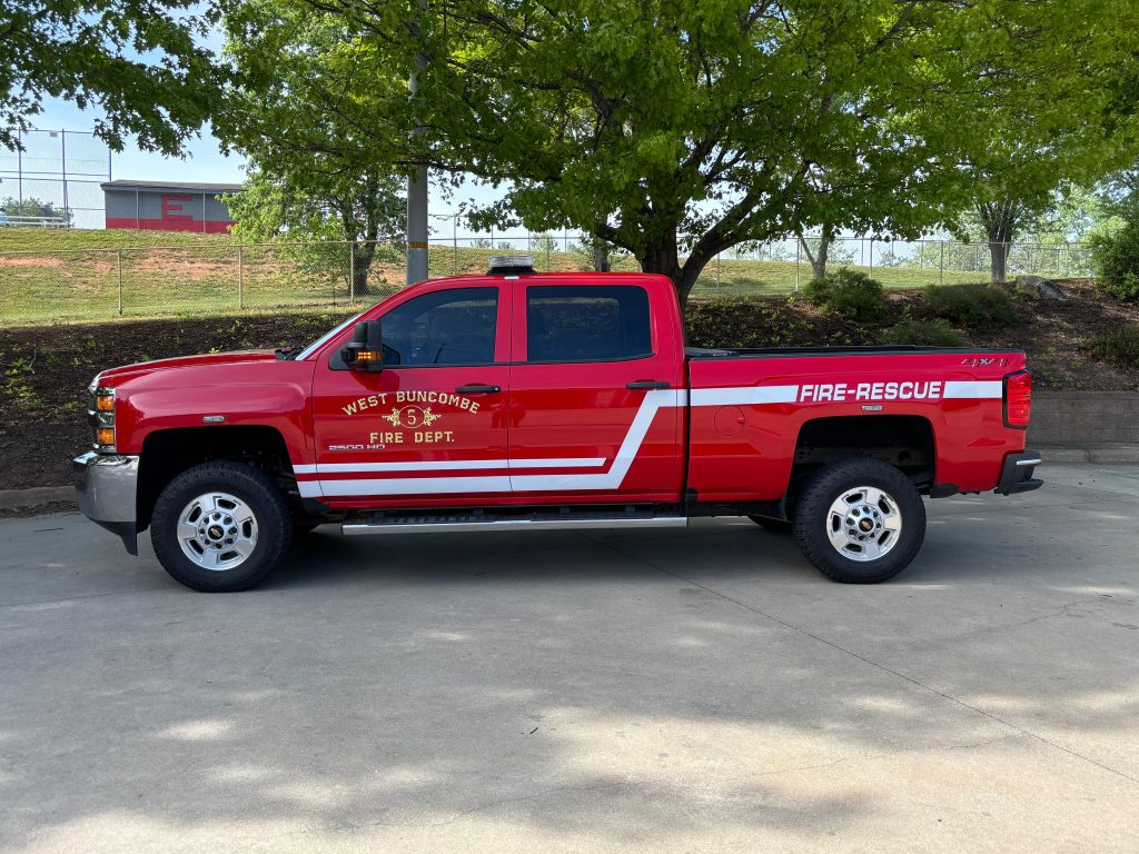 A red pickup truck with "West Buncombe Fire Dept." and "FIRE-RESCUE" written on the side is parked under a leafy tree on a sunny day.
