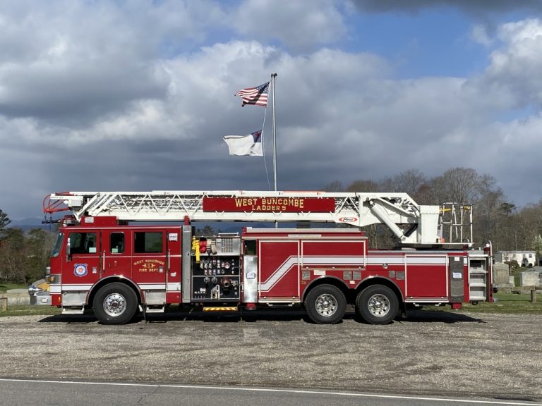 A red fire truck with "West Buncombe Ladder 9" written on the side is parked on a paved area. The truck is equipped with an extended ladder and various equipment. Two flags, one American and one white, are flown on poles in the background under a cloudy sky.