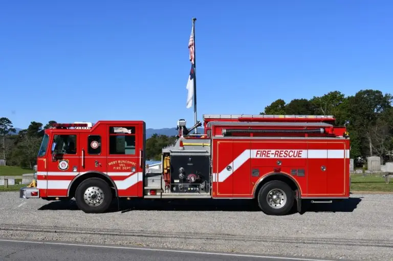 A red fire-rescue truck from the East Buncombe Fire Department is parked on a gravel surface. It displays white stripes and fire-rescue markings. The background includes trees, a flagpole with two flags, and a clear blue sky.