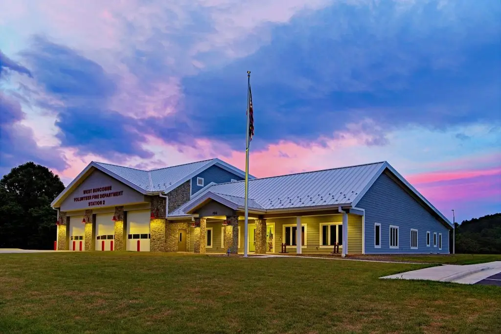 A modern fire station with a light yellow exterior and white roof under a colorful sunset sky with pink and blue hues. The building has three garage doors, a flagpole in front, and sits on a large grass lawn. Trees and a parking area are visible in the background.