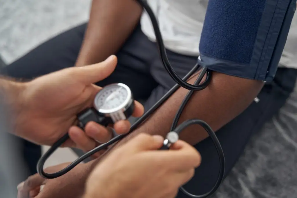 A healthcare professional measures a person's blood pressure using a sphygmomanometer. The person's arm is extended with a blue cuff wrapped around their upper arm. The healthcare professional holds the gauge and stethoscope, focusing on the measurement.