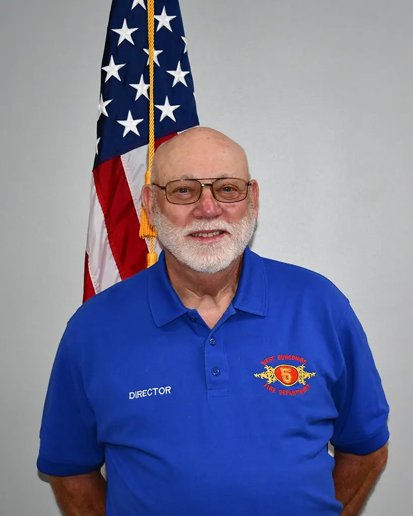 A man with a white beard and wearing glasses stands in front of an American flag. He is wearing a blue polo shirt with the word "DIRECTOR" and a fire department emblem embroidered on it.
