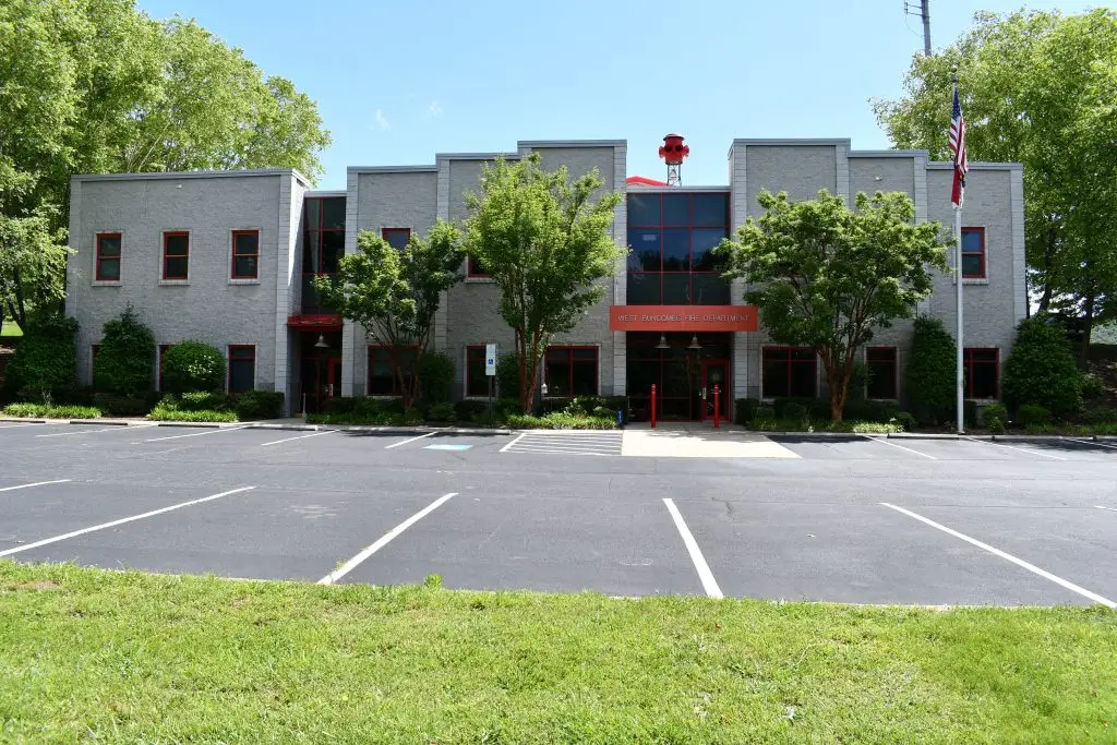 A two-story gray brick building with red-framed windows and doors, surrounded by trees. An American flag is on a pole to the right. The building's front entrance is accessible by a paved pathway, with a mostly empty parking lot in the foreground.