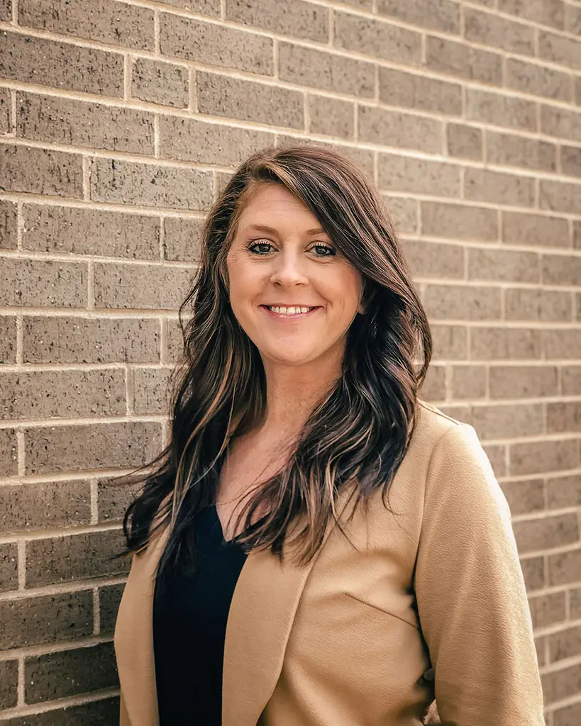 A woman with long, dark hair smiles at the camera while standing against a brick wall. She is wearing a tan blazer over a black top.