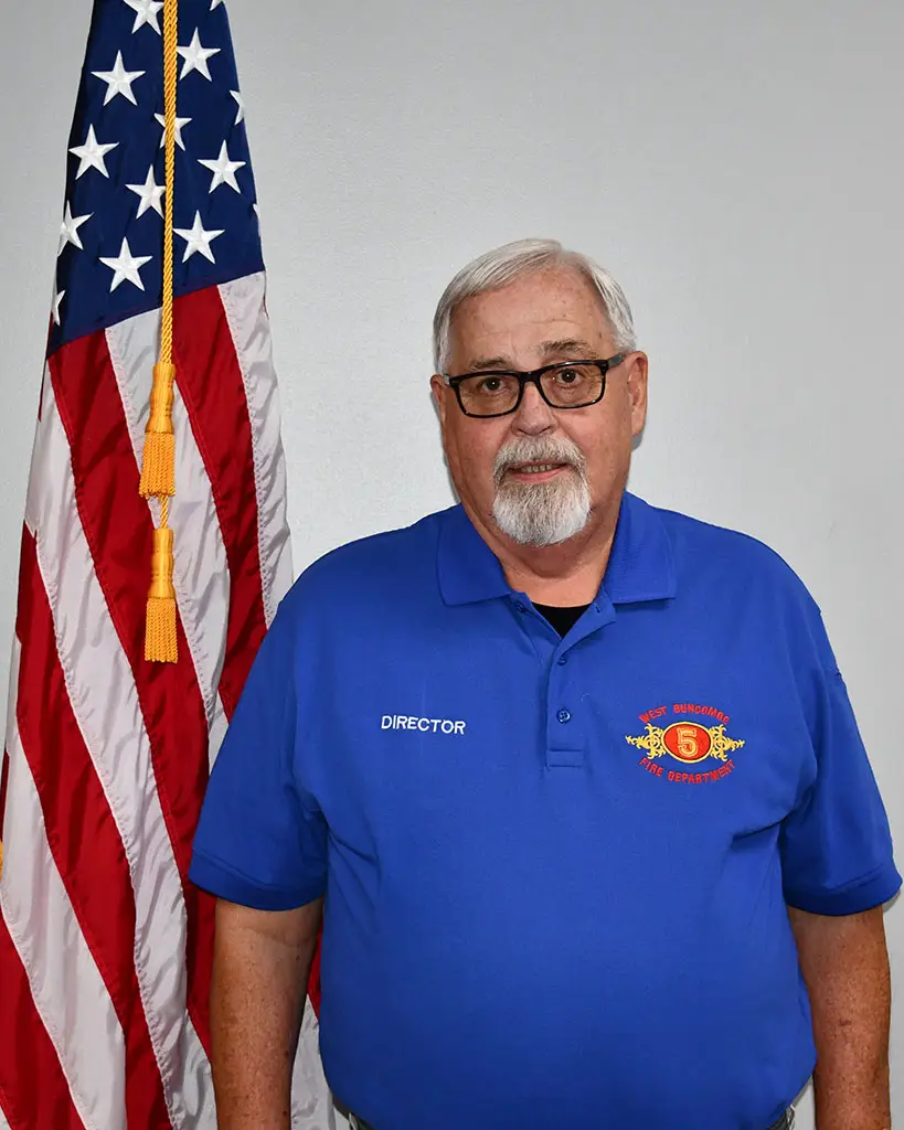 An older man with white hair and a beard is standing next to the American flag. He is wearing glasses and a blue polo shirt with an embroidered logo and the word "DIRECTOR" on it. The background is plain.