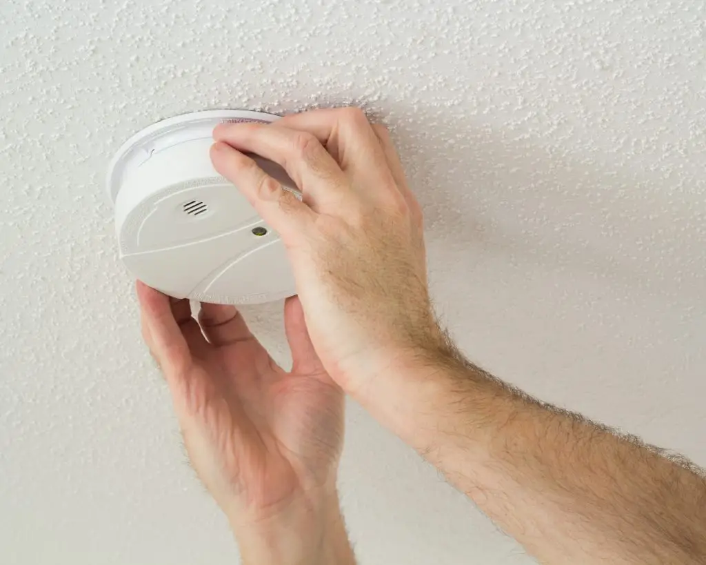 A person is installing a smoke detector on a white-textured ceiling. Both hands are securing the rounded, white device into position. The smoke detector has a small circular vent and an LED indicator light.