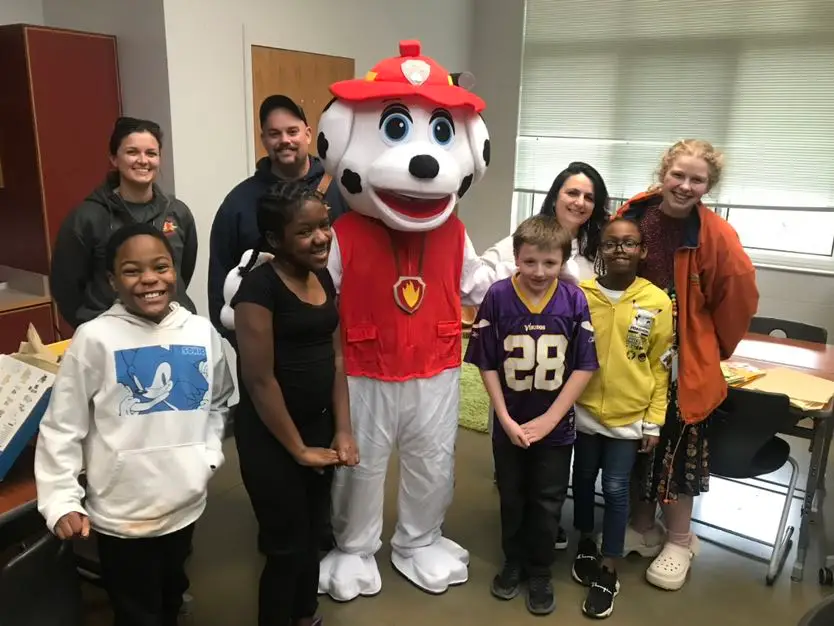 A group of children and adults pose indoors with someone dressed in a large, plush Dalmatian mascot costume wearing a red firefighter outfit. The children are smiling, and a classroom setting is visible in the background.