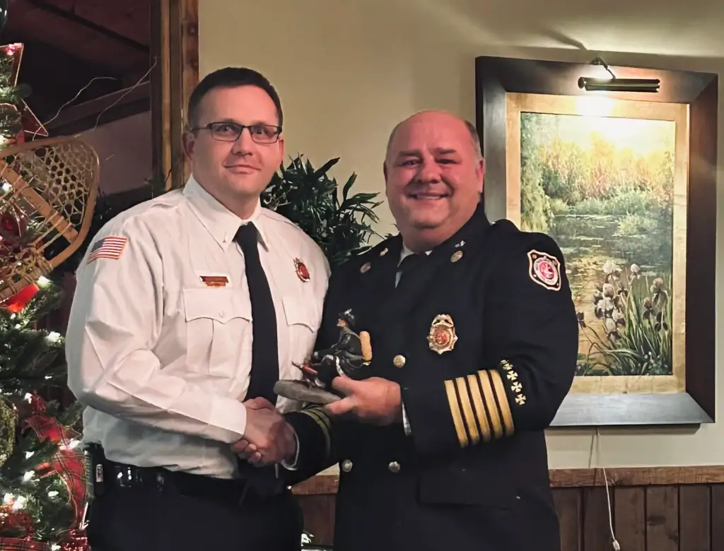 Two men in formal fire department uniforms shake hands and smile. One holds a small firefighter statue award. They stand in a room with a Christmas tree in the background, along with a framed nature painting on the wall.