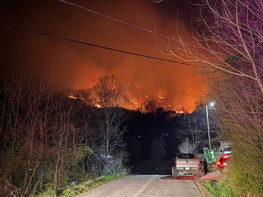 A large wildfire burns intensely on a hillside at night, illuminating the sky with orange flames. Trees border a road in the foreground where a pickup truck and car are parked, lights on. Dark, smoky sky and silhouettes of trees are visible.