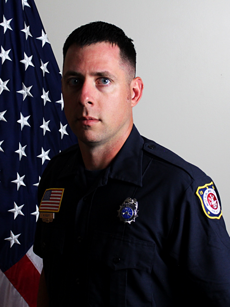 A man in a dark uniform with badges stands in front of a U.S. flag, looking at the camera with a serious expression against a plain background.