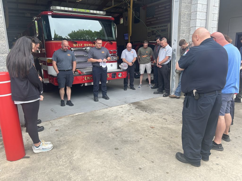 A group of firefighters and civilians stand in a circle with heads bowed in front of a fire truck inside a fire station, participating in a moment of silence or prayer.