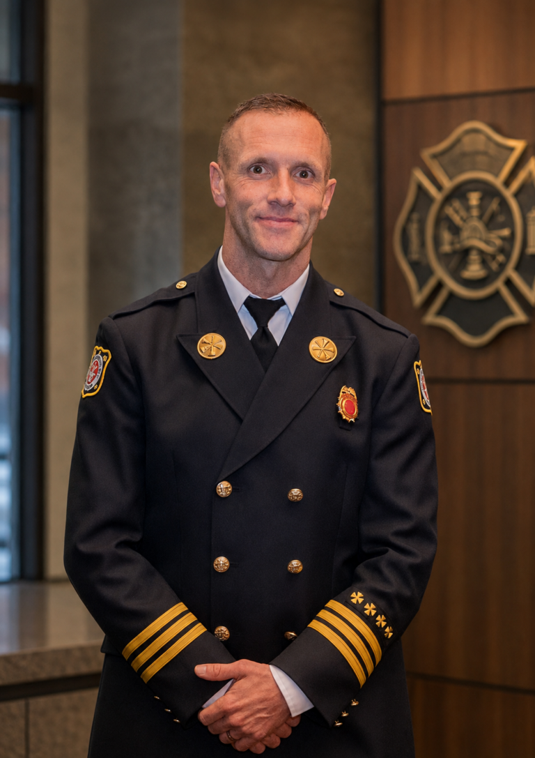 A firefighter in a formal dress uniform stands indoors, hands clasped, with a fire department emblem on the wall behind him.